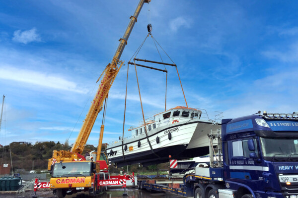 A mobile crane lifts a small boat onto the back of a transportation lorry