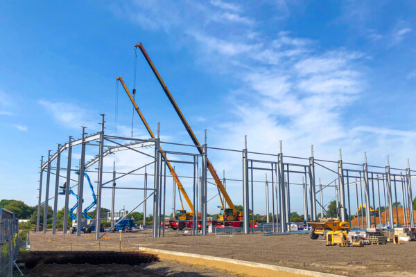 Two mobile cranes work in tandem in the distance within a steel frame of a warehouse