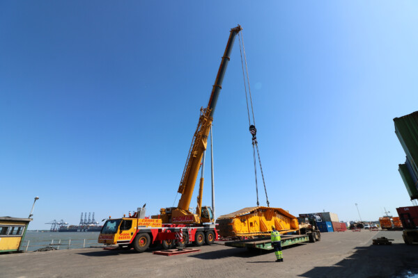 Yellow mobile crane works in a port, lifting a large piece of machinery