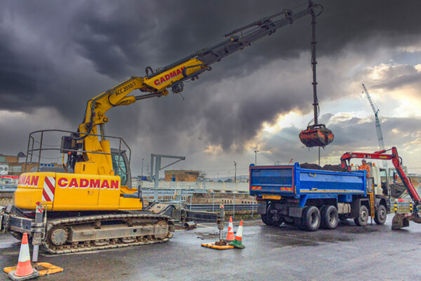 A compact crawler crane loads a transportation lorry with waste material from a grab