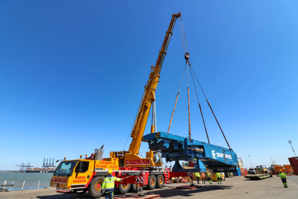 A large mobile crane lifts a piece of heavy machinery at a port with blue sky in the background