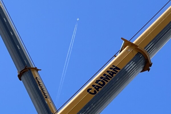 a plane flies between two mobile crane booms against a blue sky