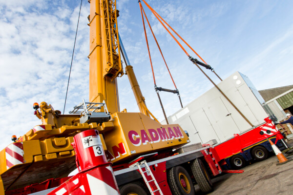 a yellow mobile crane lifts an acoustic enclosure using specialist lifting beams