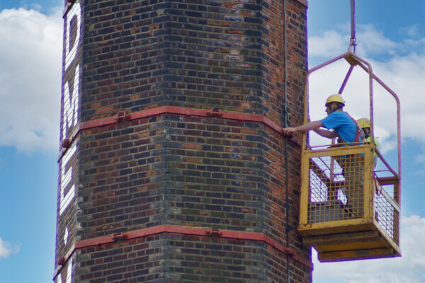 Two men work to restore a large chimney whilst inside a man cage suspended from a mobile crane