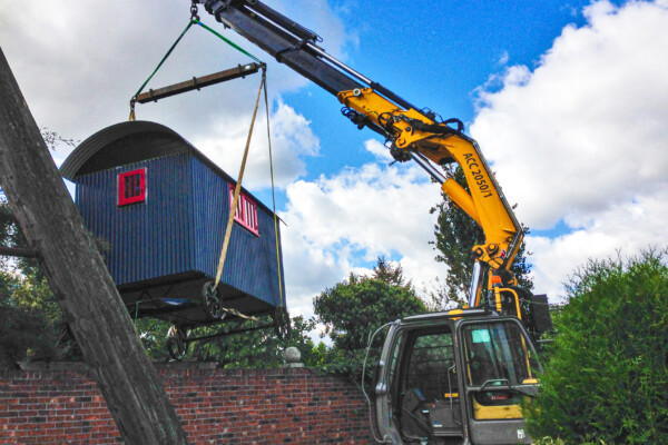 A shepherds hut is lifted over a red brick wall with a compact crawler crane