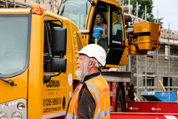 two construction workers in white hard hats are pictured next to a mobile crane. One of them is operating the crane.