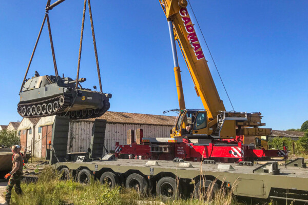 A mobile crane suspends an army tank in the air as it positions it onto the back of a transport lorry