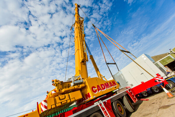 Yellow mobile crane lifts acoustic enclosure from transport lorry using specialist lifting beams