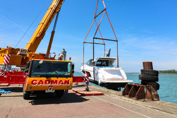A yellow mobile crane suspends a boat over the edge of marina as it lowers it into the water