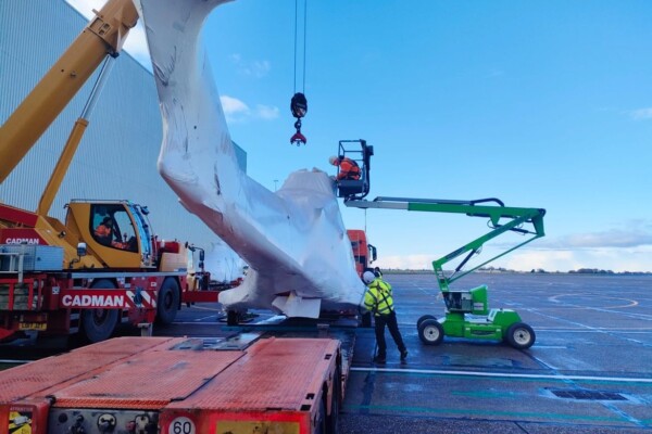 Men in hi-vis clothing inspect helicopter wrapped in plastic whilst a crane hook hangs above awaiting connection