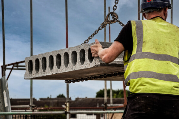 construction worker stands on site holding hollowcore plank as it is installed with a crane